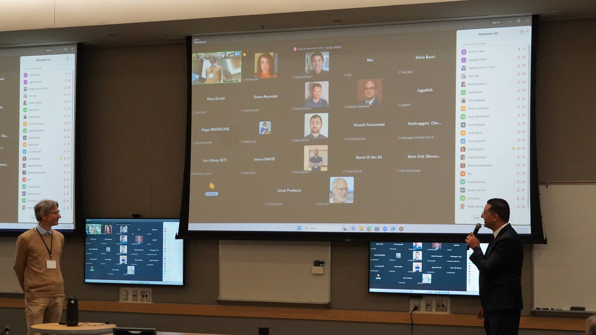 Dean, Marouane Kessentini presenters stand at the front of a lecture room while a hybrid conference session takes place. A large screen shows participants joining via Zoom, and smaller monitors display the same meeting interface